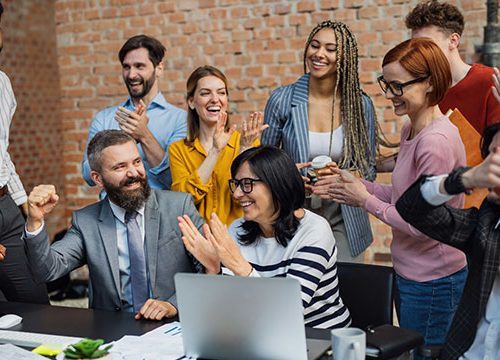 Mixed group of men and woman stood clapping and celebrating in an office with a brick wall background.
