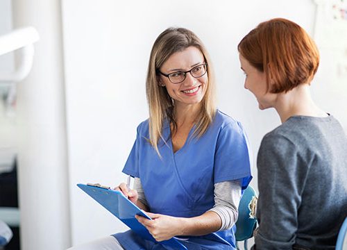Female, blonde nurse with a clipboard in her hands talking to her female patient with short read hair.