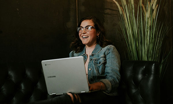 A young woman wearing glasses and smiling, sat on a black leather sofa with a laptop in her lap.