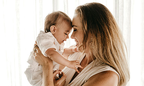 A woman with long blonde hair cuddles her baby in front of a white curtain.