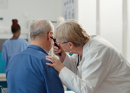 An older female health professional bending down towards an older mans ear with a temperature taker.