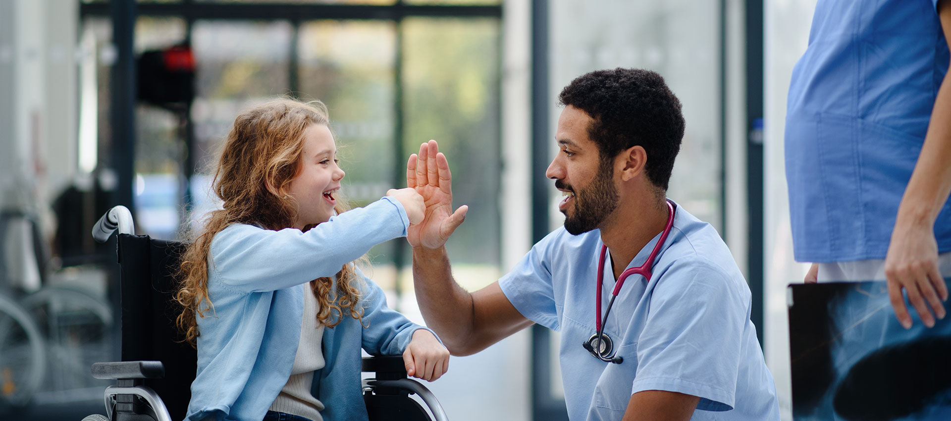 A male nurse with dark hair and dark facial hair squats down next to a little girl in a wheelchair to give her a high five.