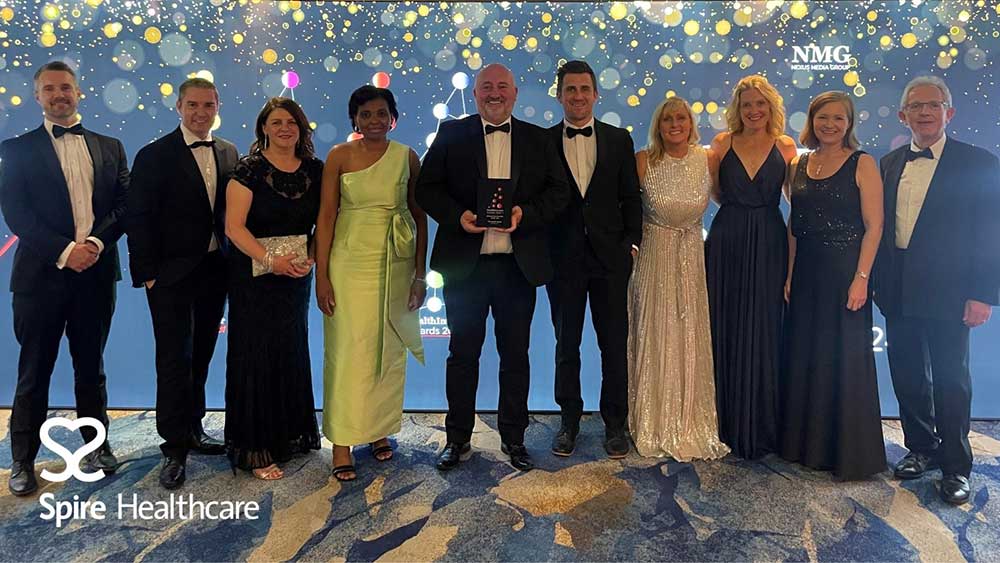 A group of ten people, dressed formally, stand together smiling. One person holds an award. The background features a blue and gold design with text: "Spire Healthcare," "NMG," and "Healthcare Awards 2023."