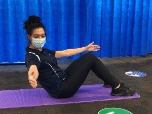 A person in black exercise attire performs an abdominal exercise on a purple yoga mat, balancing while leaning back, in a room with blue curtains. They wear a face mask.
