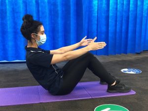 A person exercises on a purple yoga mat, performing a sit-up. They wear a face mask and black athletic clothing, with a blue curtain backdrop and gym equipment nearby.
