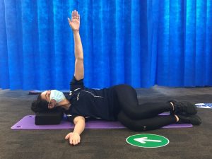 A person in a mask lies on their side on a mat, raising one arm vertically. A blue curtain forms the background. A green circular floor sign shows a white arrow.