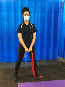 A person in athletic attire uses a red resistance band for exercise in front of bright blue curtains, standing on a black carpeted floor.