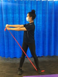 A person exercises by stretching a red resistance band horizontally with both hands. They stand on a purple mat, wearing a mask, in front of a bright blue curtain.