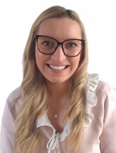 A person with glasses and long blonde hair smiles warmly, wearing a light blouse with ruffled edges, set against a plain white background.