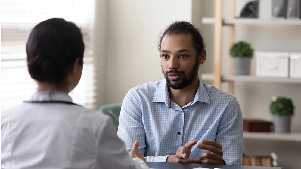A healthcare professional and a patient are seated and conversing in a well-lit office with shelves containing books and plants, emphasizing a professional healthcare consultation environment.