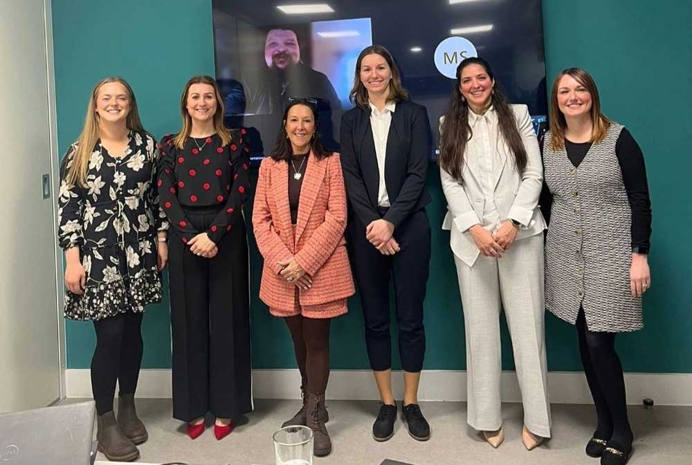 Six women stand smiling in a conference room, dressed in professional attire. A video call participant is visible on a screen behind them on a teal wall, creating a collaborative atmosphere.