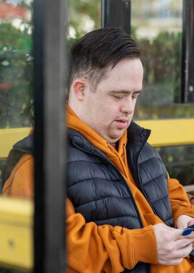 A person in an orange hoodie and black vest sits at a bus stop, focused on a smartphone. The background shows foliage through the shelter's glass panels.