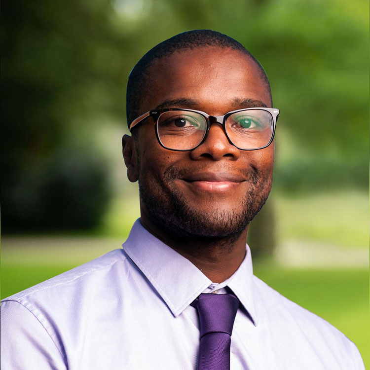 A person in a white shirt and purple tie stands outdoors, smiling. The background features green foliage and grass, suggesting a park or garden setting.