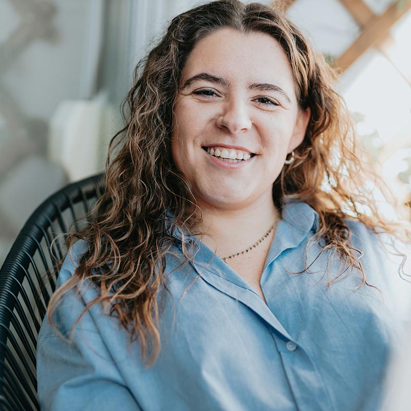 A person is smiling while sitting indoors, wearing a blue shirt. They appear relaxed and approachable, conveying a sense of warmth and friendliness in a professional setting.