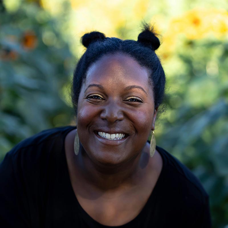 A person smiles broadly, standing outdoors with a blurred background of green foliage and hints of yellow, possibly flowers, in a sunlit setting.