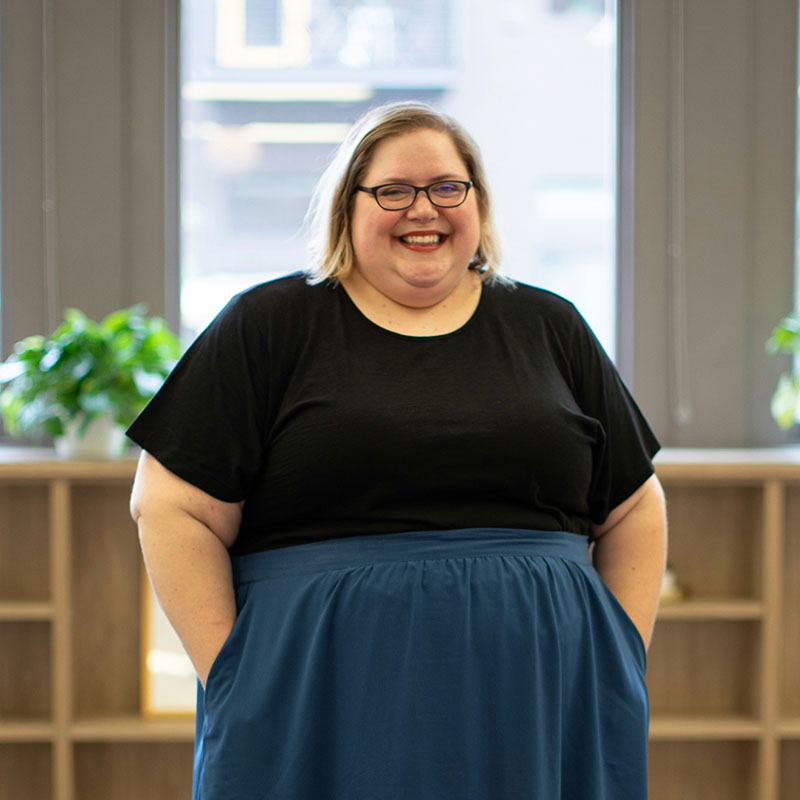 A person is standing and smiling, wearing glasses, a black top, and a blue skirt. They are in an indoor setting with shelves and plants in the background.