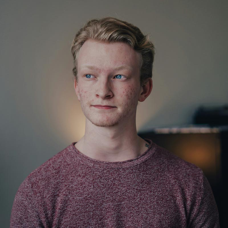 A person with light hair, wearing a maroon sweater, looks to the side in a softly lit room with a blurred background.