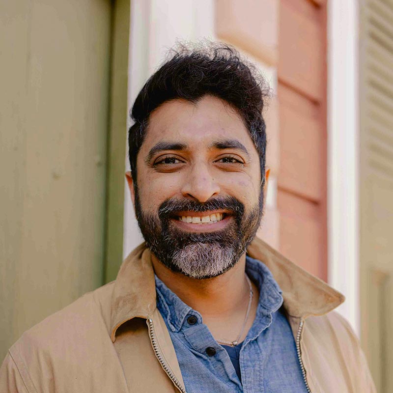 A man smiles warmly, wearing a blue shirt and tan jacket, standing outdoors against a background of colorful building siding.