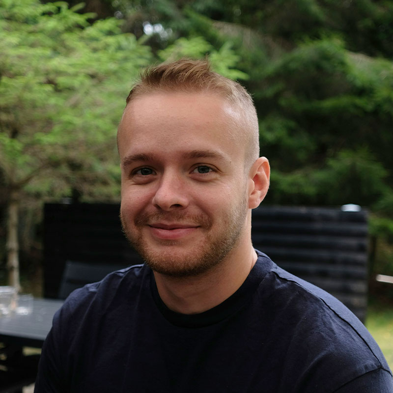 A person is sitting outdoors, smiling slightly at the camera. The background features lush greenery, creating a serene, natural setting.