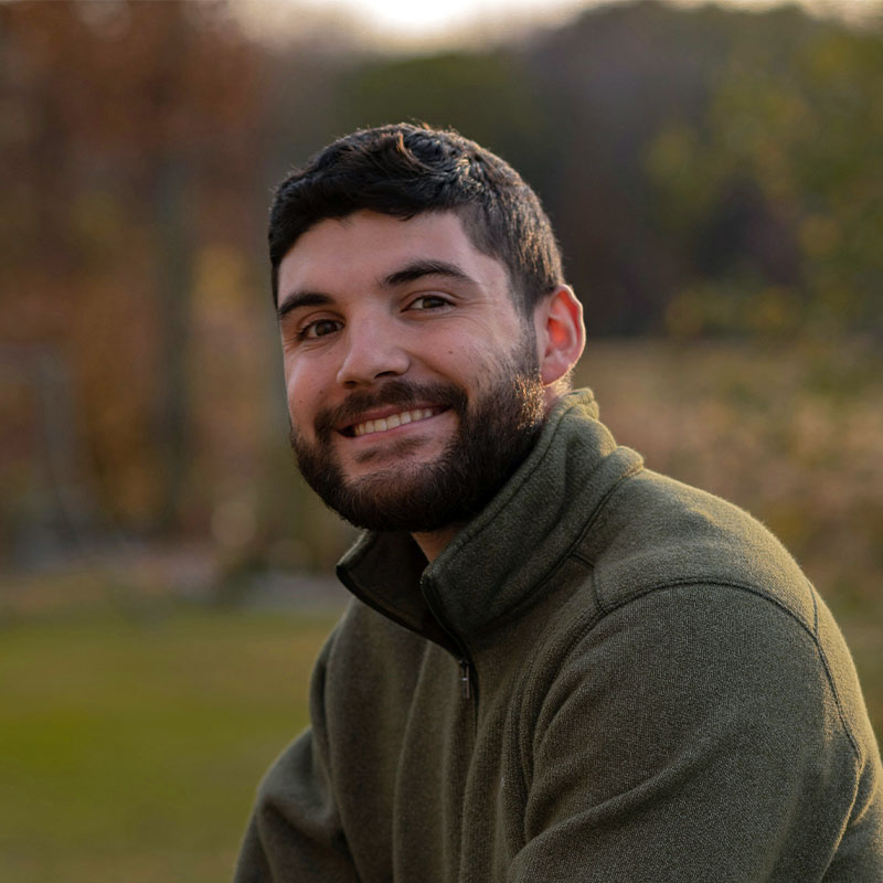A man wearing a dark green zip up fleece smiles at the camera while stood in a park.