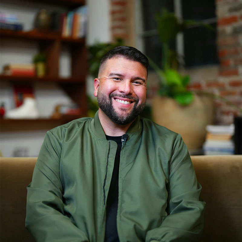 A person is sitting and smiling on a sofa. They are wearing a green jacket. The background features a bookshelf with books and plants, and a brick wall.