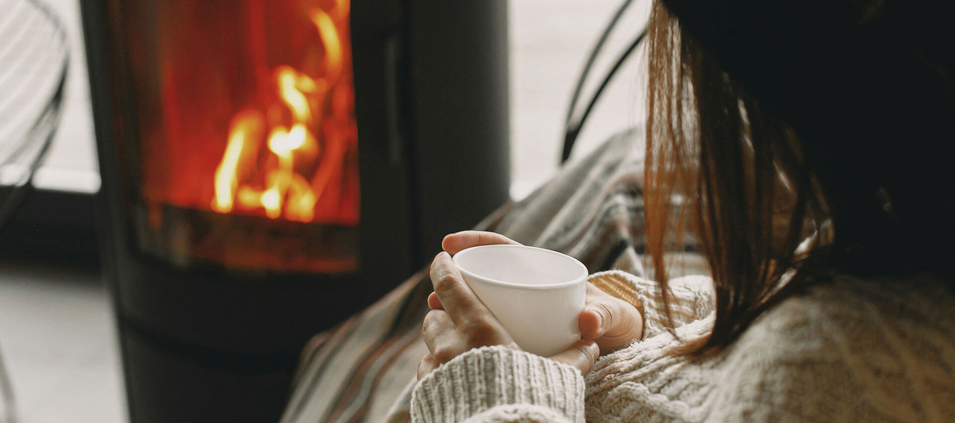 A close up of a woman with shoulder length brown hair holding a white cup sitting in front of a log burner with roaring flames.