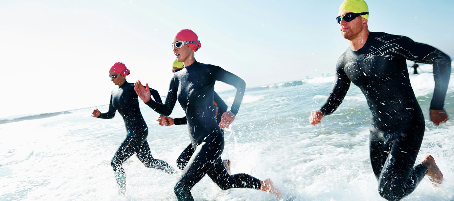 A man and 2 women in black wetsuits and pink and yellow swimming caps and goggles, run through shallow waves in a cold sea.