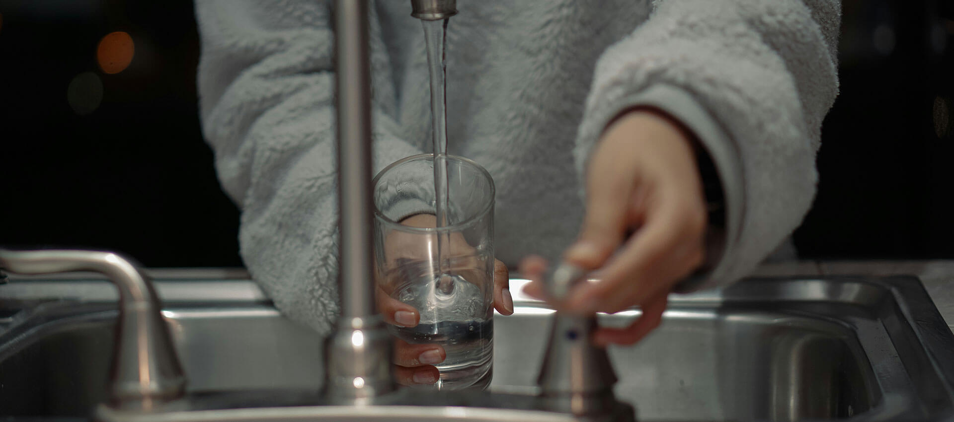 A hand pours clear liquid from a jug into a glass on a kitchen countertop, surrounded by other kitchenware.