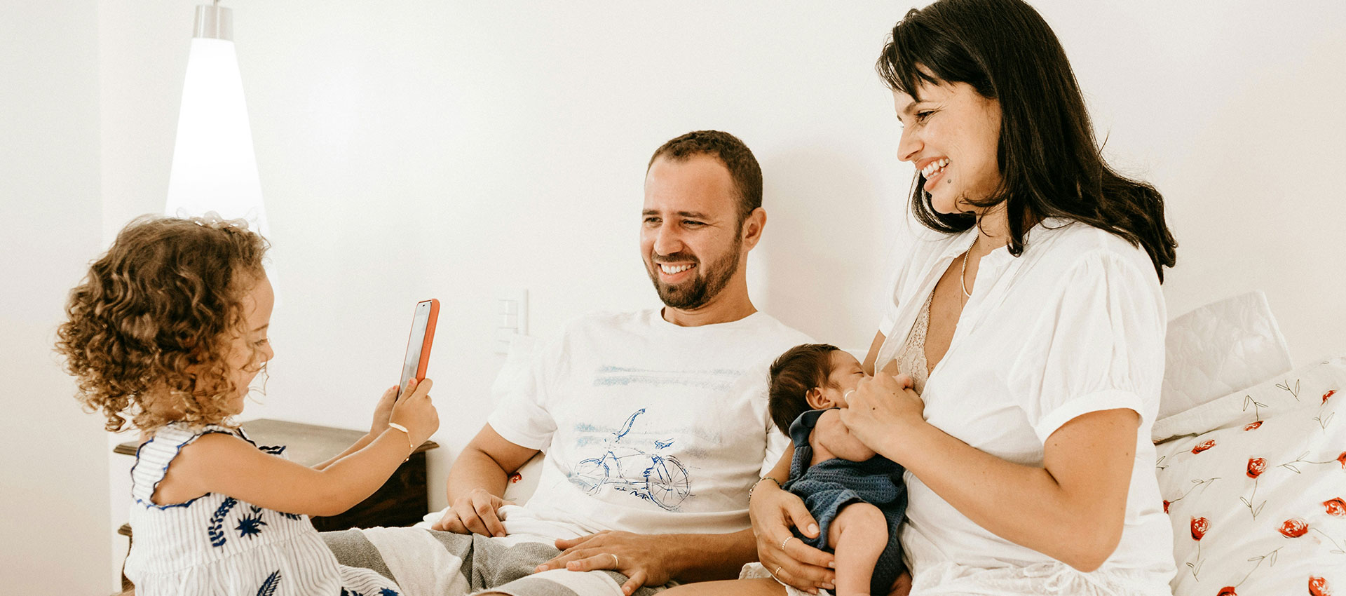 A family including a man, woman, little girl and a newborn baby sit on bed laughing.