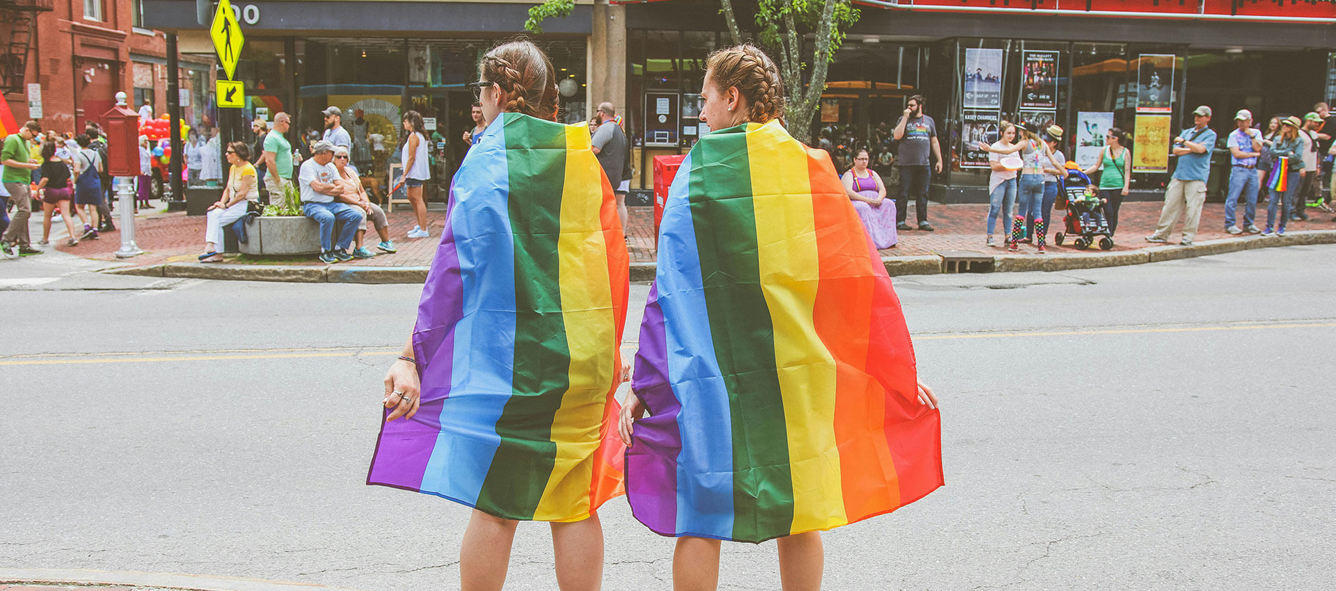 Two people draped in rainbow flags stand on a street, observing a crowd on the opposite sidewalk at a Pride event.