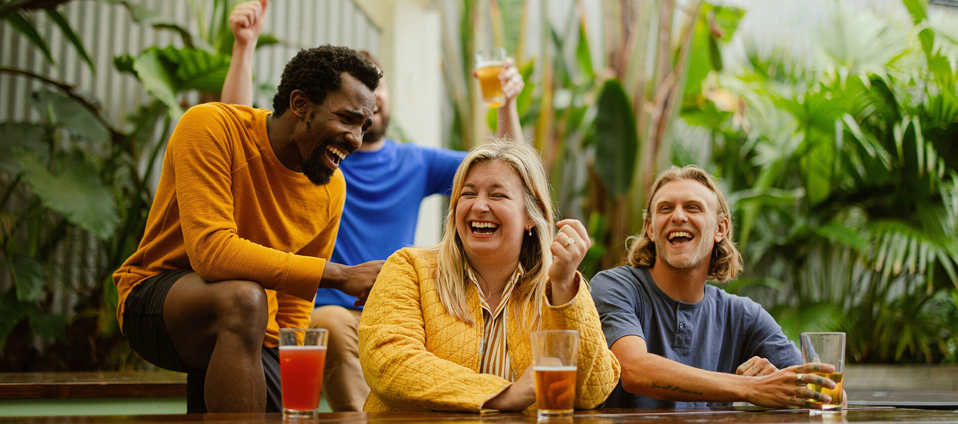 A group of people laugh and enjoy drinks at a wooden table, surrounded by lush greenery. One person holds a drink in the background, creating a lively, joyful atmosphere.