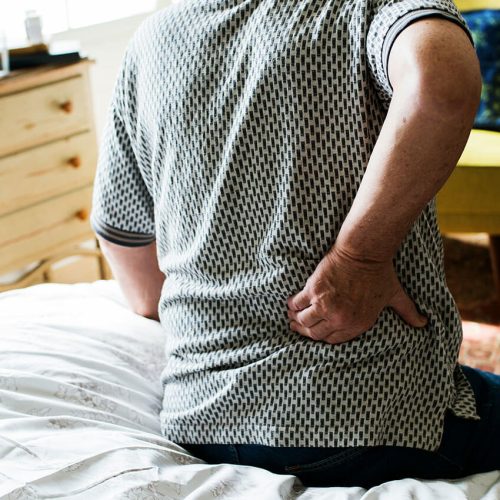 A man in a patterned polo top sits on the end of a bed in rubbing his lower back to ease pain.