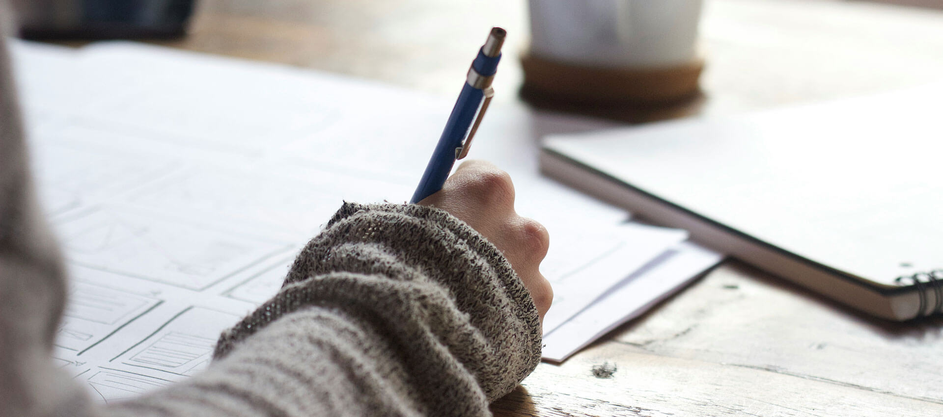 A close up of a girl with a pen in her hand drawing on white paper at a wooden deak.