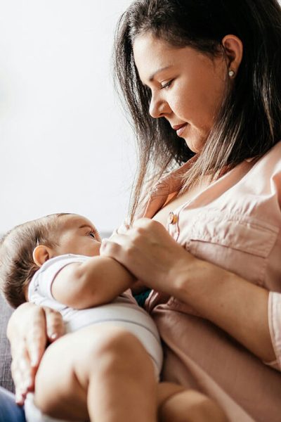 A person feeds a baby with a bottle while sitting on a beige couch. A floor lamp and a small table with decorations are in the background.