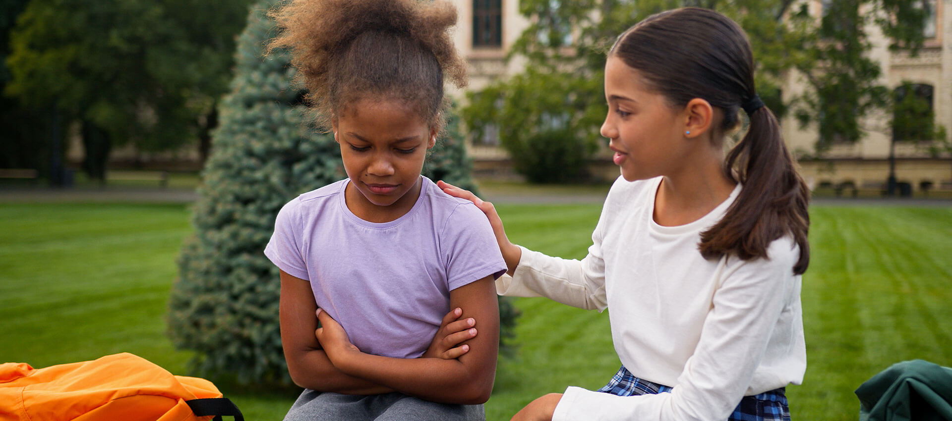 A girl in a purple shirt sits with arms crossed, looking down. Another girl in white places a comforting hand on her back. They are outdoors near a tree in a park setting.