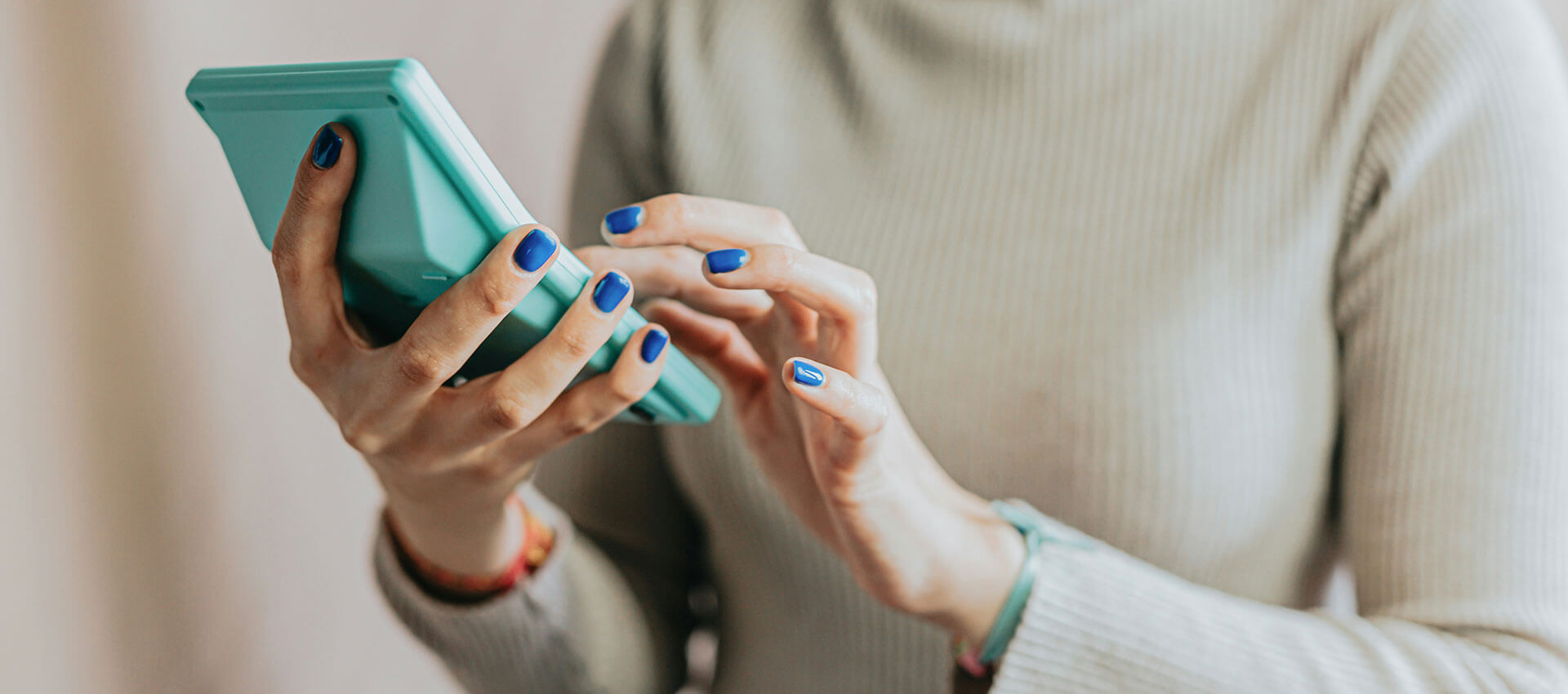 A person holds a green smartphone and types with one hand in a light-colored sweater, focusing on the phone screen in a casual indoor setting.