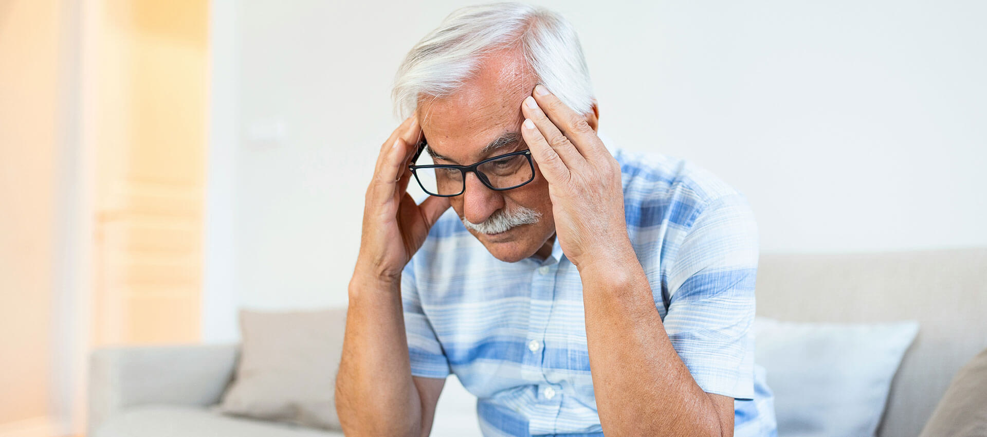 An older man with grey hair and black glasses, sits on a cream sofa with his head in hands.