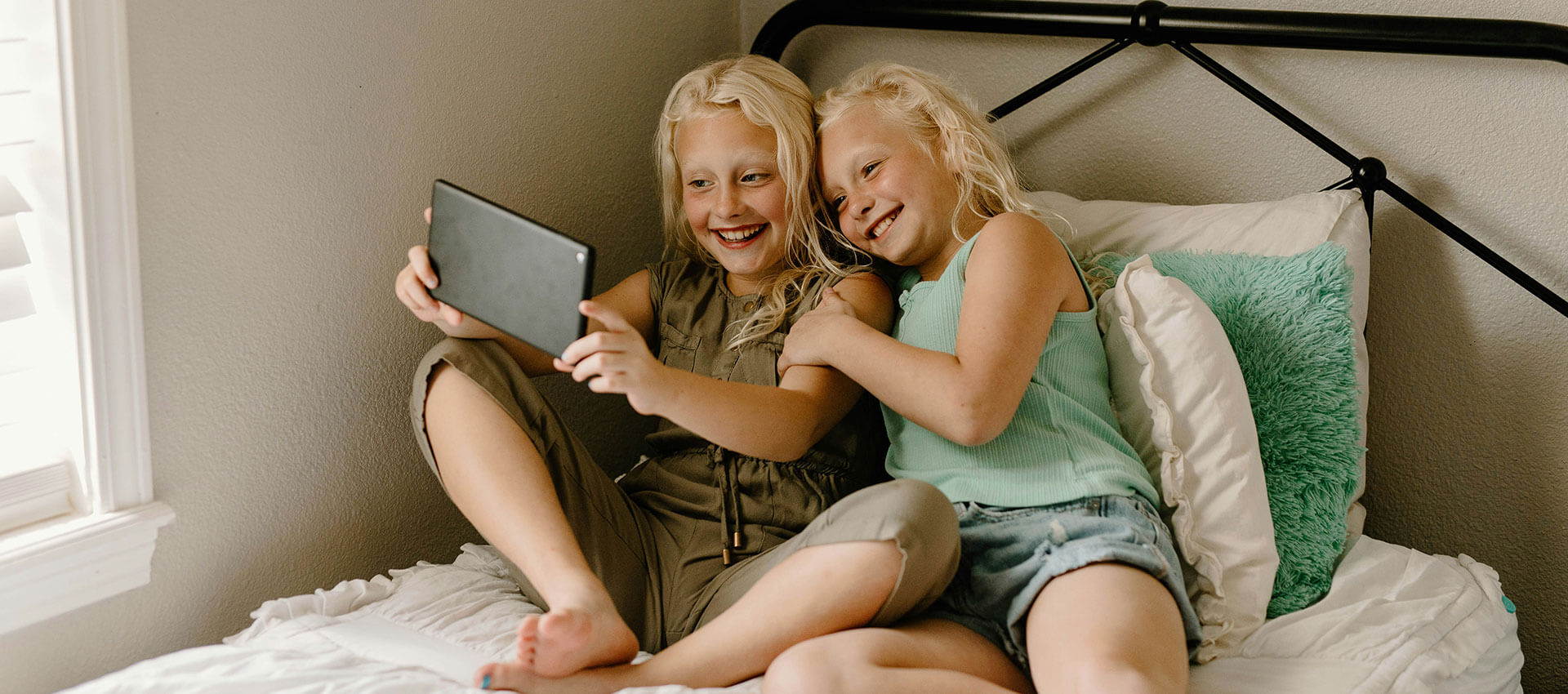 Two young girls with long blonde hair sit on a bed taking pictures of themselves on a tablet device.