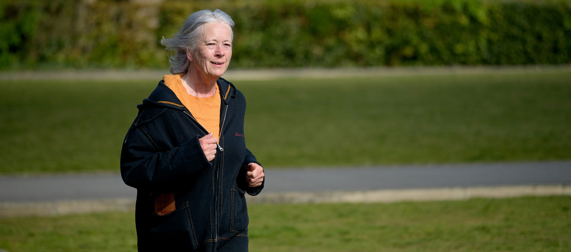 An elderly person jogs on a grassy field, wearing a black jacket over an orange shirt. A path and green hedge are visible in the background.