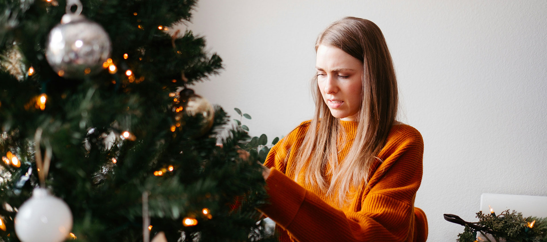 A young woman with long hair, wearing an orange jumper stands at a Christmas tree adding decorations.