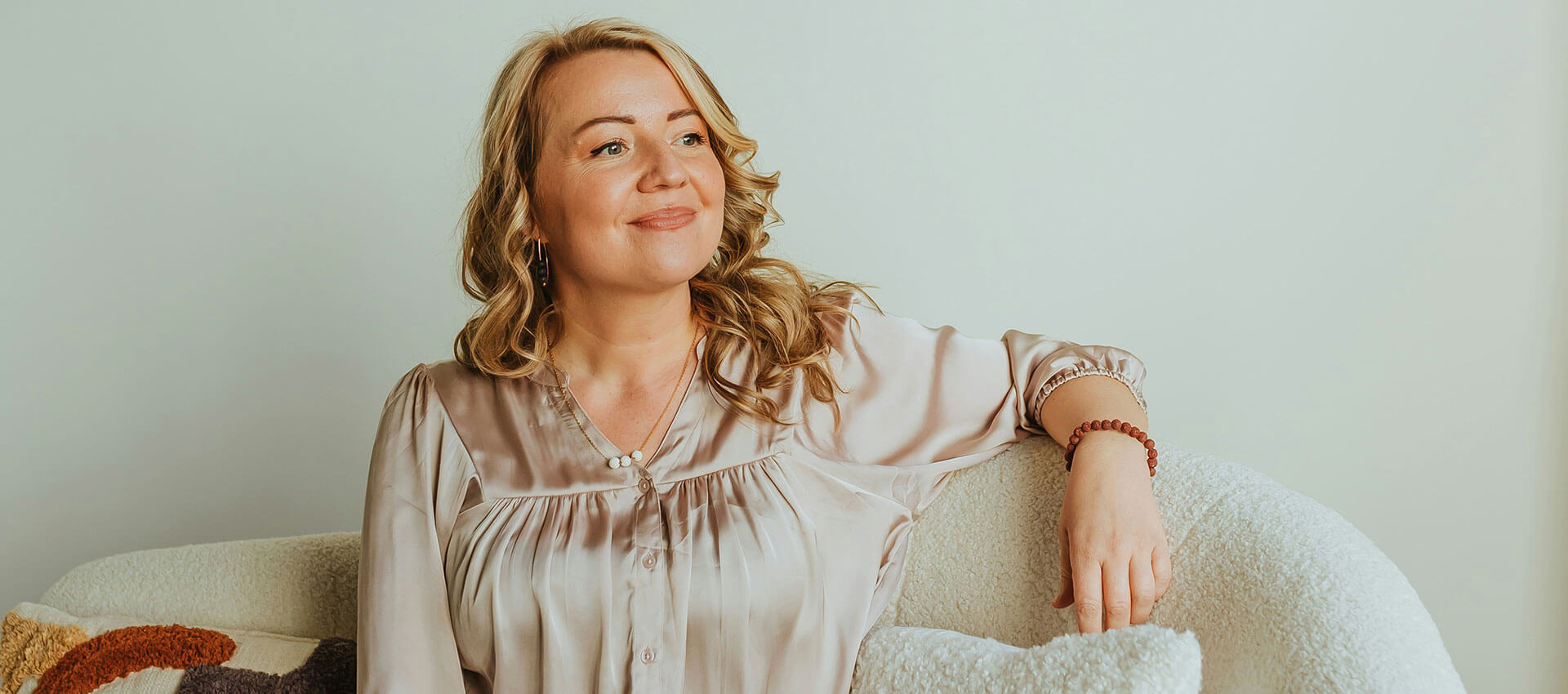 A person sits comfortably on a sofa, smiling with one arm resting on the backrest. The setting includes a cozy, neutral-toned room with a textured cushion beside them.