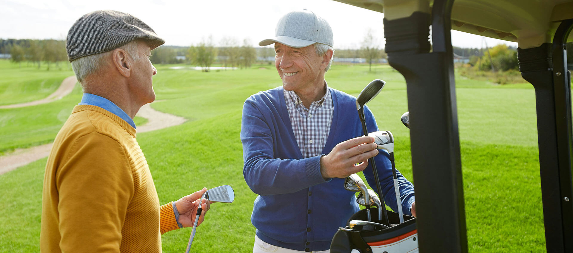 Two elderly men, one in a flat cap and orange sweater, and the other in a baseball cap and blue cardigan, stand by a golf cart smiling on a golf course.