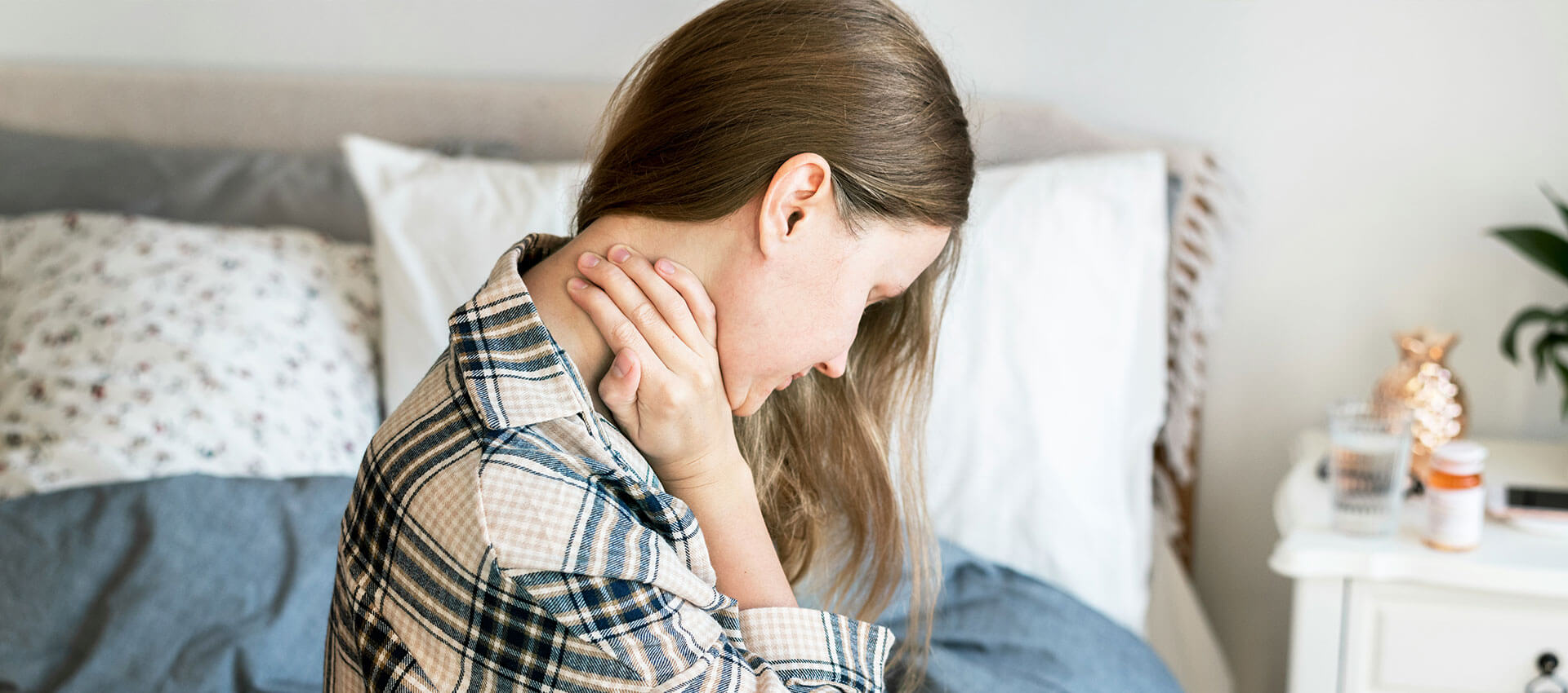 A person holding their neck in discomfort sits on a bed, surrounded by pillows and a bedside table with a glass, pill bottle, and plant.