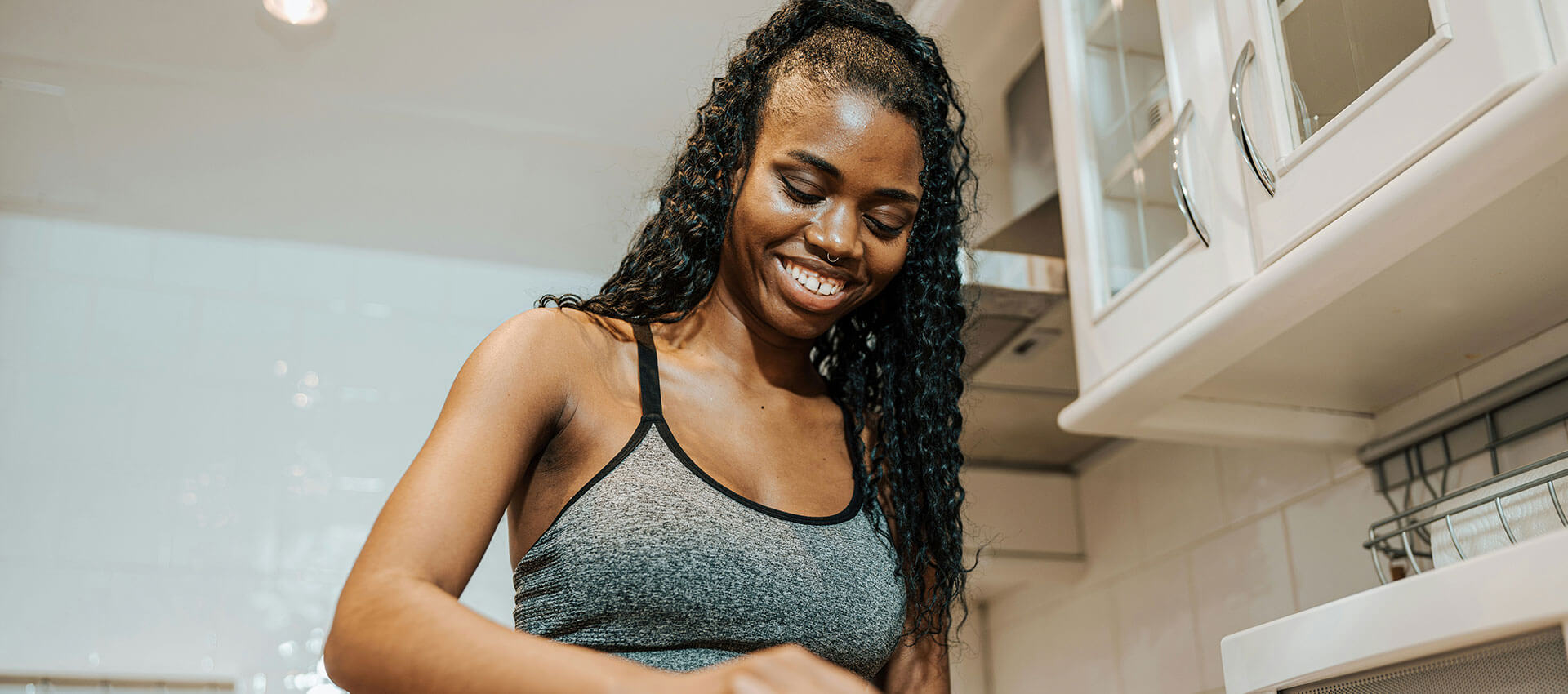 A black woman with long dark hair wearing a grey sport bra looks down at her hands in a cream kitchen.