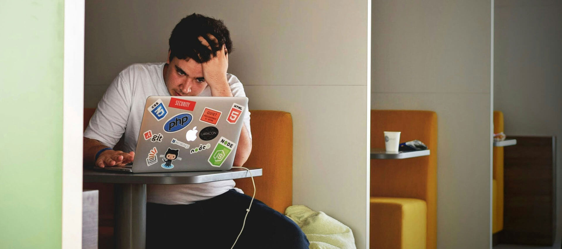 A man sits in a meeting pod in a modern office building with a stressed expression on his face and his hand running through his hair while looking at a silver laptop covered in colourful stickers.