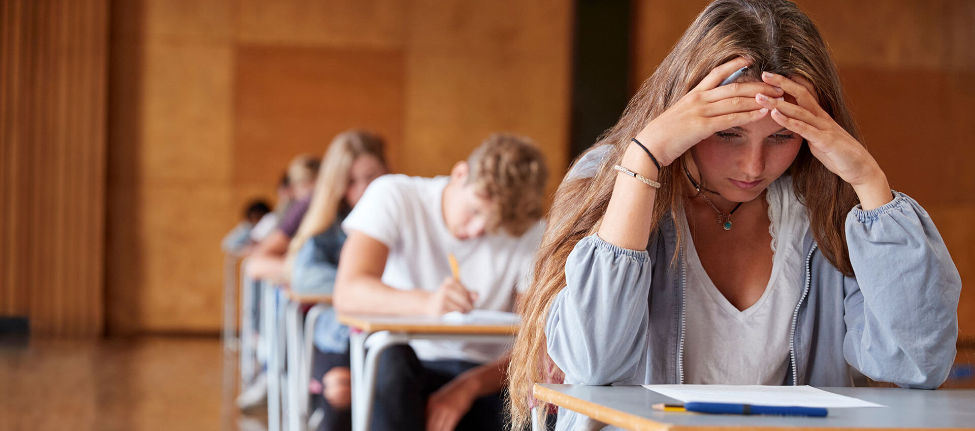 A person sits at a desk, holding their head in concentration, surrounded by others taking an exam in a large, wooden-floored room.