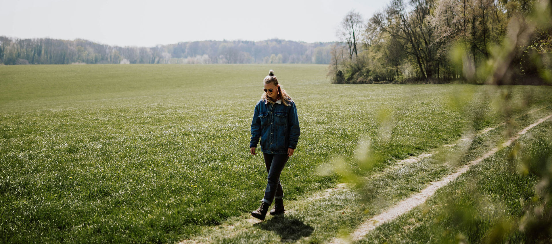A person in a denim jacket walks along a grassy path in a vast green field, with trees and distant hills in the background.