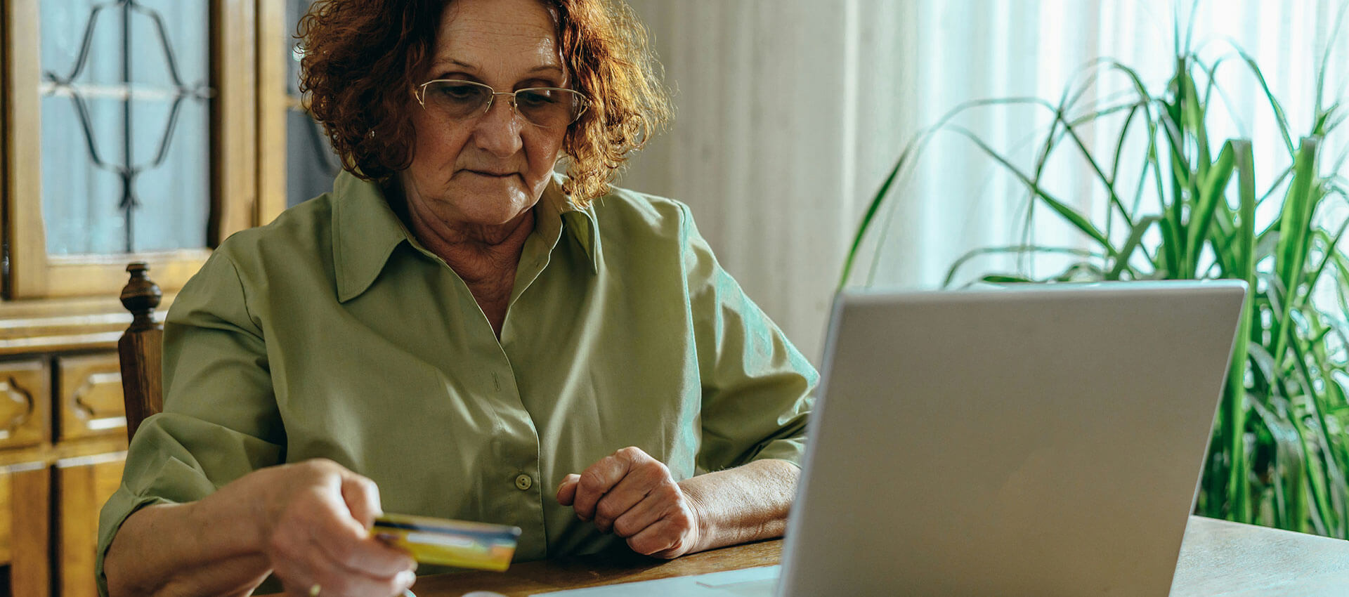 An older woman wearing glasses sits at a wooden table, holding a credit card while using a laptop. Sunlight filters through curtains in a room adorned with a tall plant.
