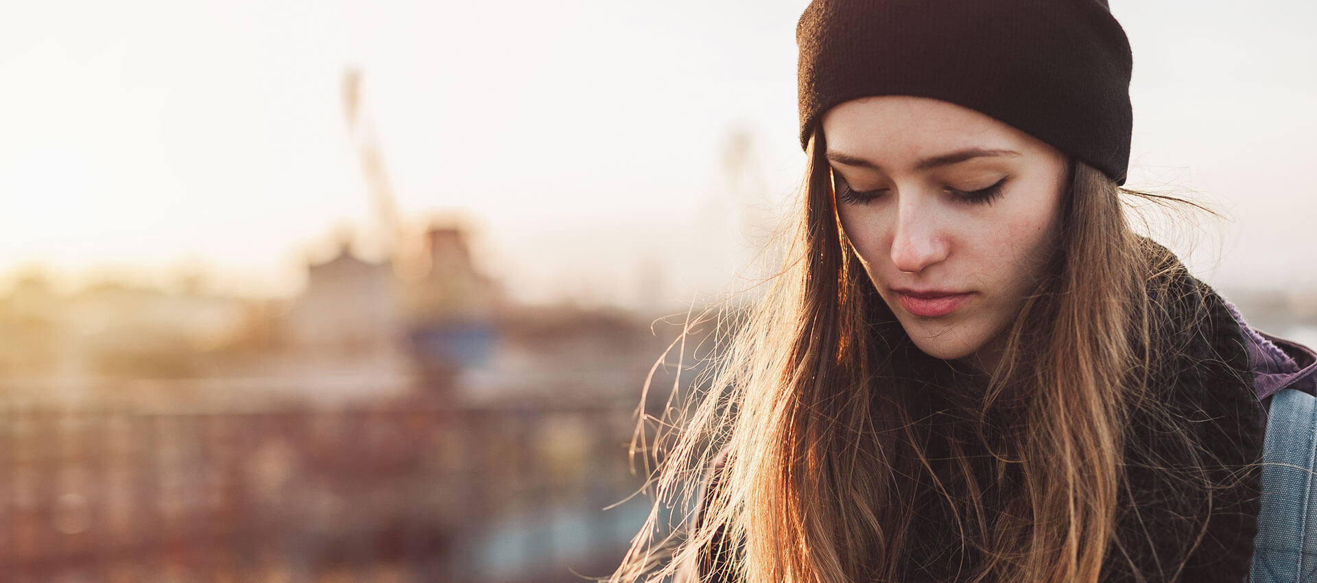 A person wearing a beanie and sweater gazes downward thoughtfully, seated outdoors with a blurred cityscape and sunset in the background.