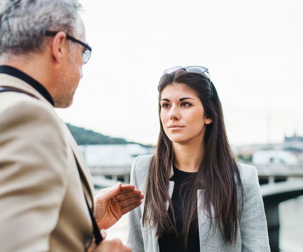 A young woman with long dark hair looks intensely at a middle aged man with grey hair and glasses outside in a busy city.
